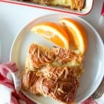 A white plate with two slices of ham and swiss croissant breakfast casserole and two orange wedges, next to a red and white cloth napkin.