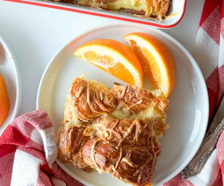 A white plate with two slices of ham and swiss croissant breakfast casserole and two orange wedges, next to a red and white cloth napkin.