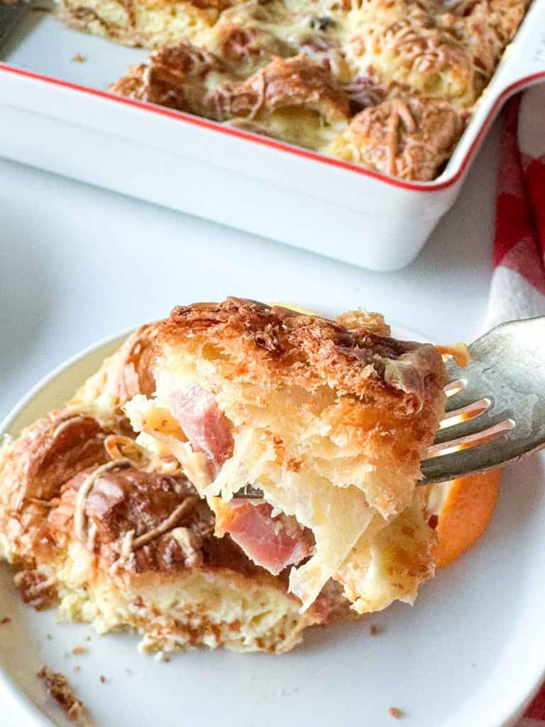 A fork holds a piece of ham and swiss croissant breakfast casserole with layers of pastry, ham, and melted cheese above a plate, with the casserole dish in the background.