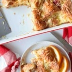 A plate with a serving of ham and swiss croissant breakfast casserole and orange slices sits next to a baking dish holding the remaining casserole, with a red and white napkin nearby.