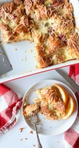 A plate with a serving of ham and swiss croissant breakfast casserole and orange slices sits next to a baking dish holding the remaining casserole, with a red and white napkin nearby.