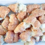 Several ham and swiss croissant pieces are torn and arranged closely together on a rectangular white tray, ready for a delicious breakfast casserole.