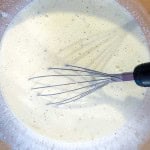 A metal whisk in a bowl of pale, frothy batter, preparing the base for a delicious ham and swiss croissant breakfast casserole.