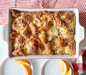 A ham and swiss croissant breakfast casserole sits baked with cheese in a rectangular dish, accompanied by two plates of orange slices on a white surface.
