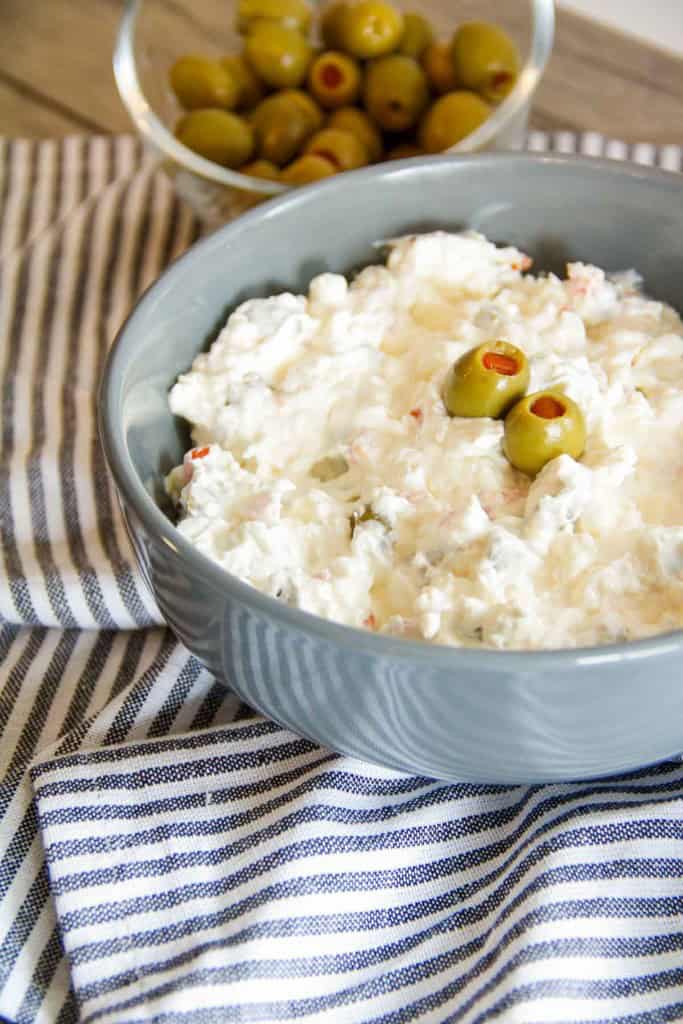 A bowl of creamy salad topped with green olives sits on a striped cloth, with a dish of green olives in the background.