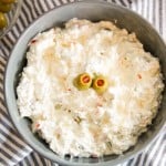 A bowl of creamy pimento cheese dip garnished with two green olives, placed on a striped cloth with a bowl of additional olives nearby.