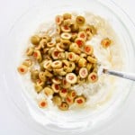 A glass bowl with a mixture of cream cheese and sliced green olives with pimentos, with a metal spoon resting inside.