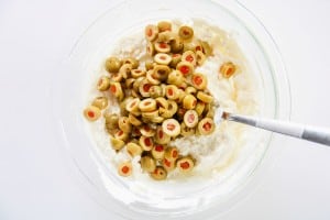 A glass bowl with a mixture of cream cheese and sliced green olives with pimentos, with a metal spoon resting inside.