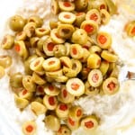 A bowl of cream cheese mixture topped with sliced green olives stuffed with pimentos, with a metal spoon on the side.