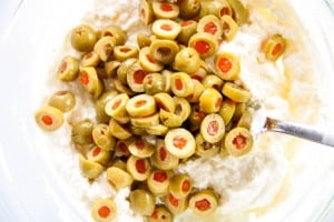A bowl of cream cheese mixture topped with sliced green olives stuffed with pimentos, with a metal spoon on the side.