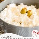 A bowl of creamy olive dip topped with green olives, placed on a table with part of an olive jar in the background.