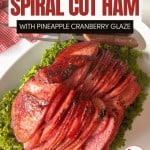 A sliced spiral-cut Baked Ham with Cranberry Pineapple Glaze is served on a bed of leafy greens.