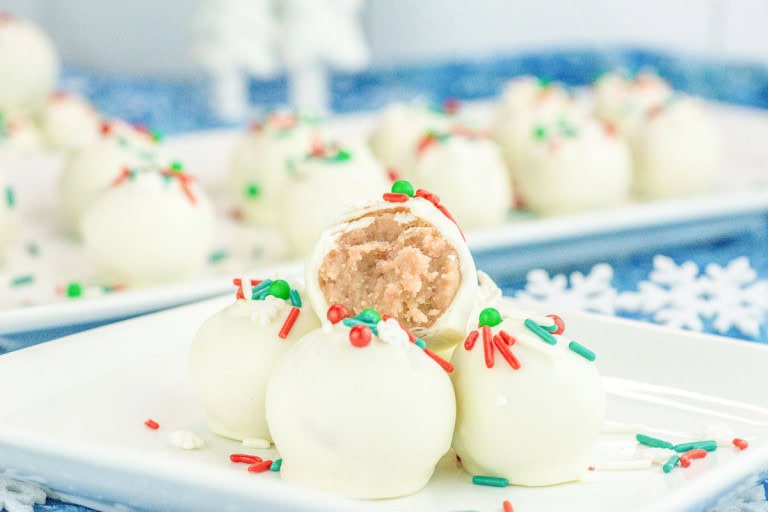 White chocolate-coated truffles topped with colorful sprinkles are arranged on a plate, with one truffle cut in half to show the creamy filling inside.