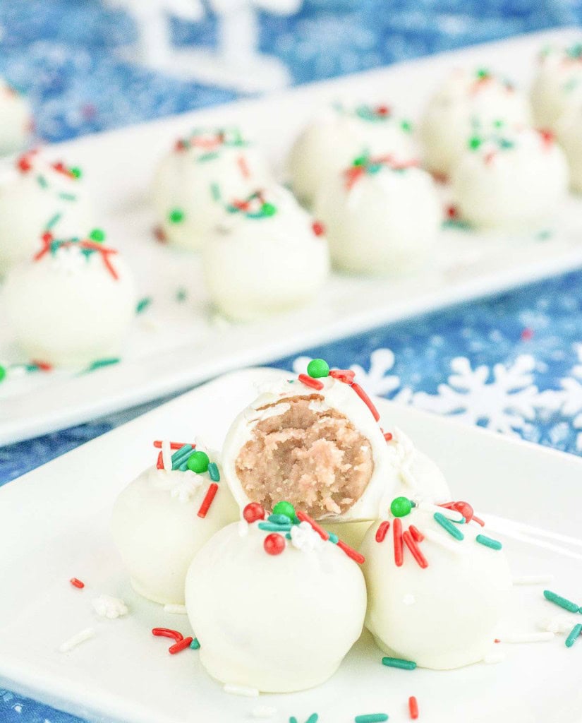 White chocolate-covered truffles with red and green sprinkles are arranged on a white plate, one cut open to show a pinkish interior. More truffles are visible on a tray in the background.