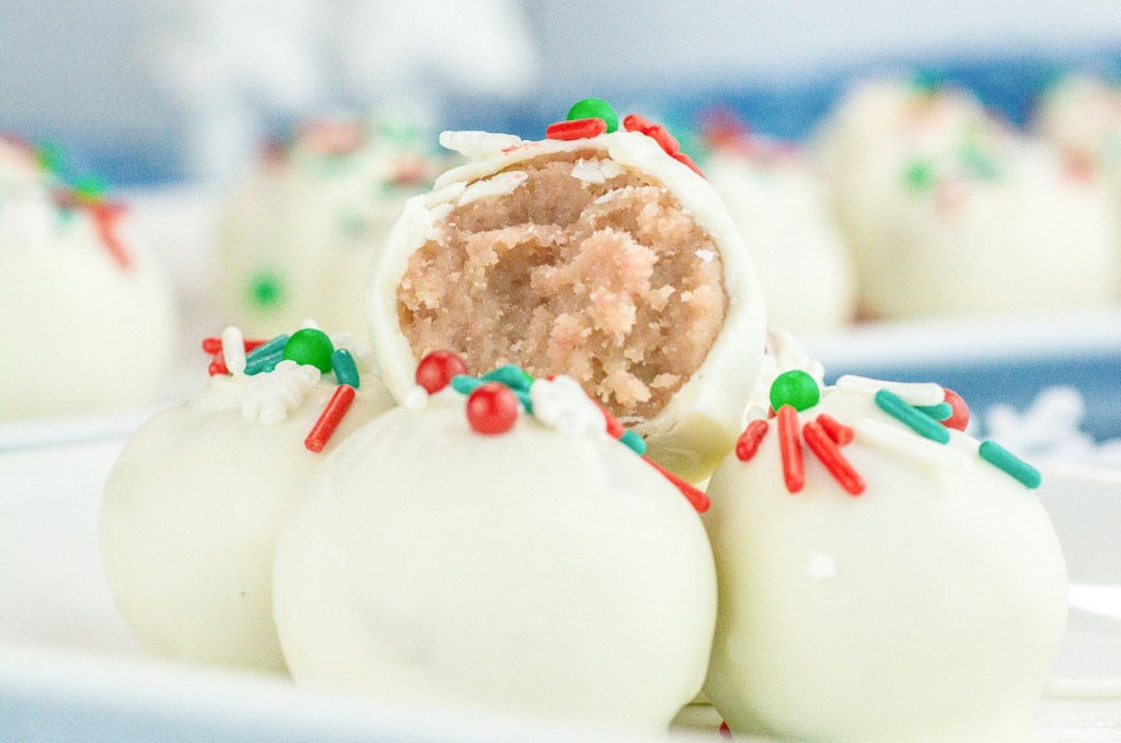 White chocolate covered truffles with red, green, and white sprinkles, one with a bite taken to show a light brown interior.