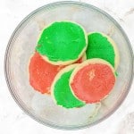 A glass bowl containing round sugar cookies with red and green frosting, placed on a white marble surface.