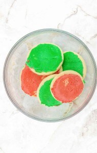 A glass bowl containing round sugar cookies with red and green frosting, placed on a white marble surface.