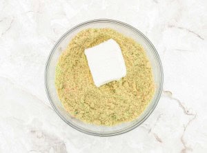 A clear glass bowl containing dry cake mix and a block of cream cheese on a light marble surface.