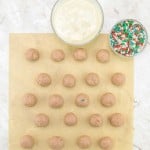 Twenty dough balls are arranged on parchment paper, with a bowl of white melted coating and a bowl of red, green, and white sprinkles nearby on a marble surface.