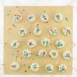 Twenty-one round, white chocolate-covered treats topped with red, green, and white sprinkles are arranged on a sheet of parchment paper.
