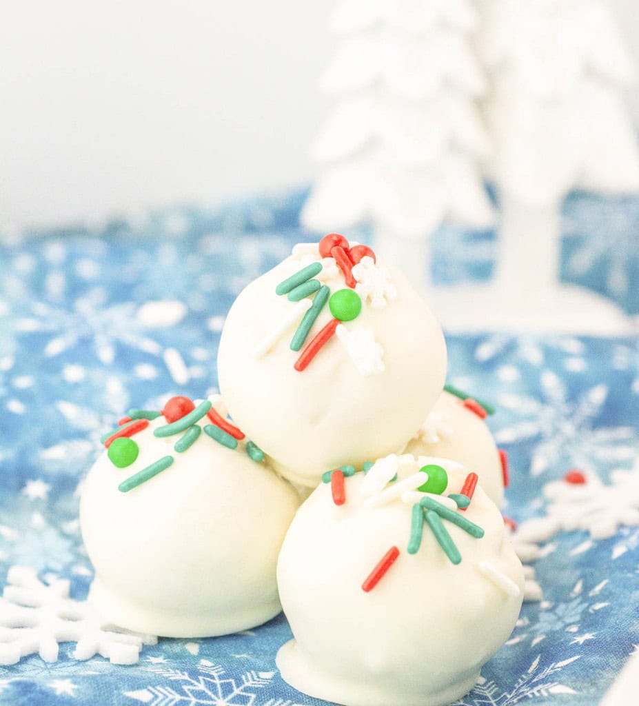 Four white chocolate-covered truffles topped with red, green, and white sprinkles are stacked on a blue snowflake-patterned cloth with white decorations in the background.