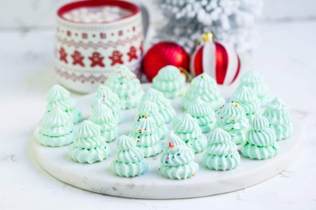 A plate of mint green meringue cookies shaped like Christmas trees, decorated with sprinkles, with holiday ornaments and a mug in the background.