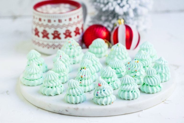 A plate of mint green meringue cookies shaped like Christmas trees, decorated with sprinkles, with holiday ornaments and a mug in the background.