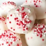 White chocolate truffles topped with red, pink, and white sprinkles are arranged on a white plate.
