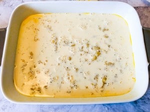 A white baking dish filled with a pale yellow, slightly bubbly liquid mixture, possibly prepared for baking.