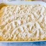 A baking dish filled with an even layer of dry, crumbly flour mixture spread over a yellow base, ready to be baked.