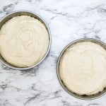 Two round cake pans filled with unbaked cake batter sit on a white marble countertop.