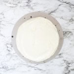 Round chantilly cake with a smooth layer of white frosting on a circular board, placed on a white marble surface.