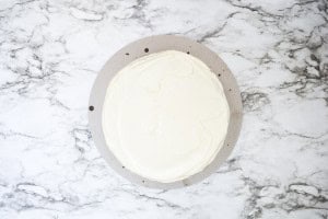 Round chantilly cake with a smooth layer of white frosting on a circular board, placed on a white marble surface.