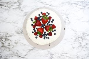 A round chantilly cake with white frosting topped with whole strawberries and blueberries, placed on a marble surface.