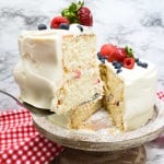 A slice of frosted chantilly cake with berries is lifted from the whole cake, which sits on a cake stand with a red-checkered cloth underneath.