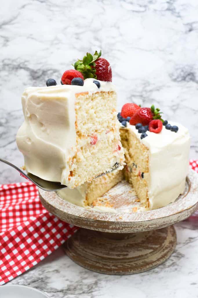 A slice of frosted chantilly cake with berries is lifted from the whole cake, which sits on a cake stand with a red-checkered cloth underneath.