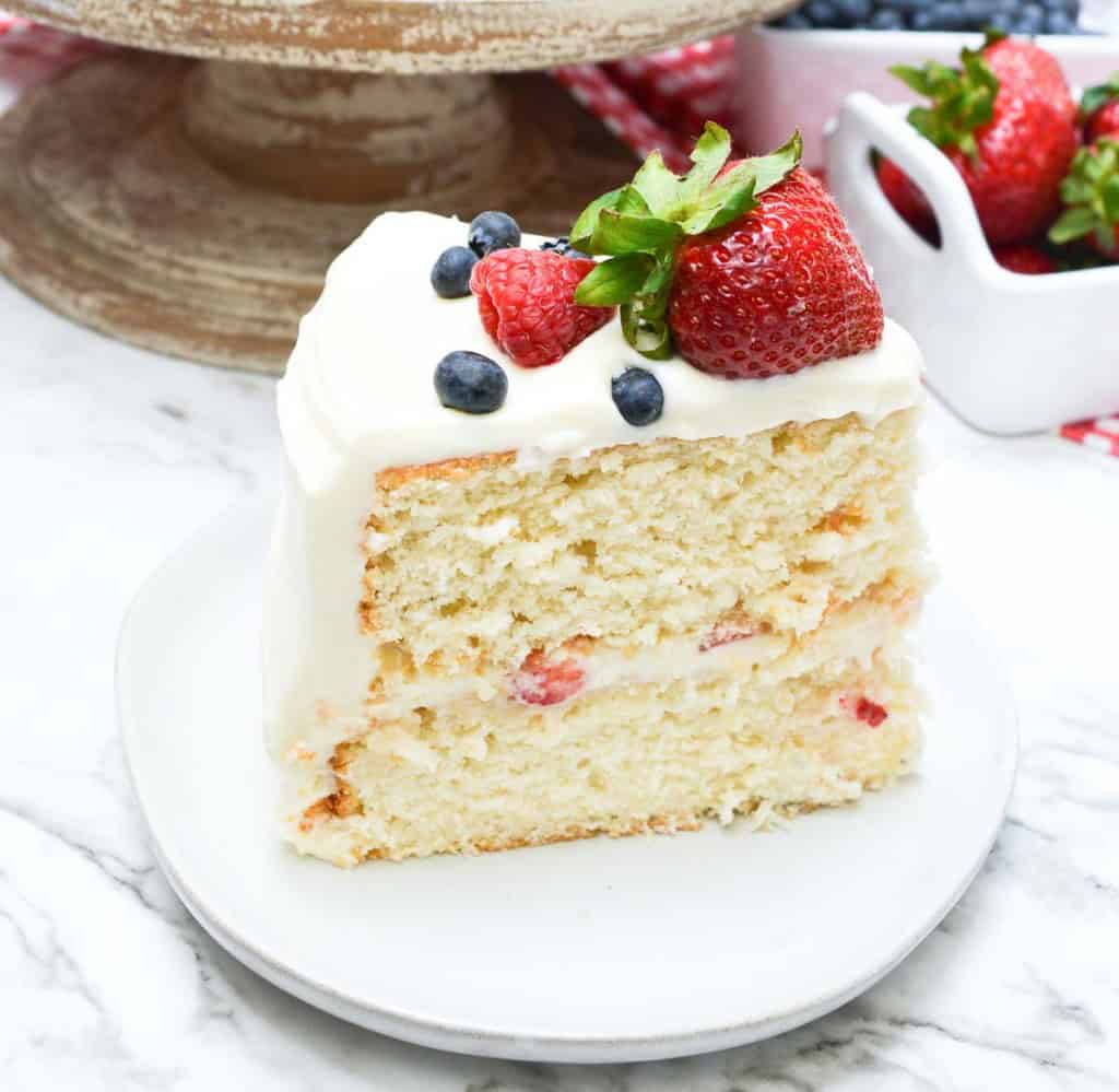 A slice of layered vanilla chantilly cake with white frosting, topped with a strawberry, blueberries, and a raspberry, served on a white plate.