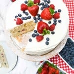 A chantilly cake frosted to perfection is topped with strawberries, raspberries, and blueberries. It sits on a stand beside bowls of fresh berries and a red checked cloth, with one slice served on a plate.