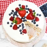 A round chantilly cake with white frosting topped with strawberries, raspberries, and blueberries, with one slice removed; bowls of berries and a red checkered cloth are nearby.