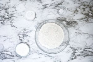 A glass bowl of flour with three small containers of white powder ingredients on a marble countertop.