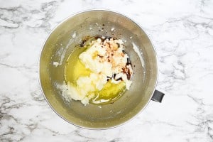 A metal mixing bowl with sugar, softened butter, eggs, and vanilla extract on a marble countertop.