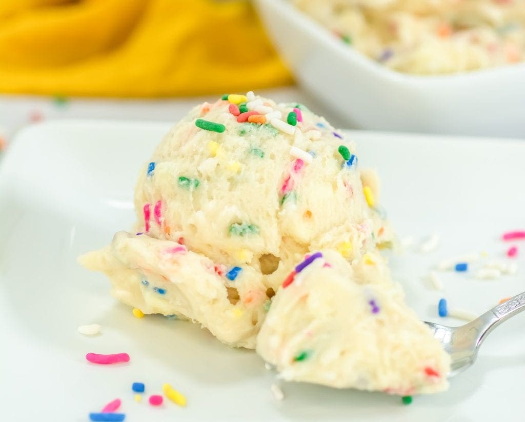A scoop of vanilla ice cream with colorful sprinkles sits on a white plate, with a spoonful taken out and more sprinkles scattered nearby.