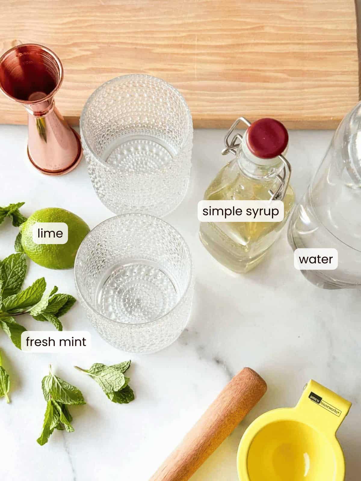 Ingredients for a mojito mocktail on a countertop: fresh mint, lime, a jigger, two glasses, simple syrup, water, a wooden muddler, and a yellow citrus juicer.