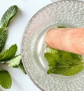 A close-up of a hand muddling fresh mint leaves in a textured glass with a muddler, with additional mint sprigs on the table beside the glass.