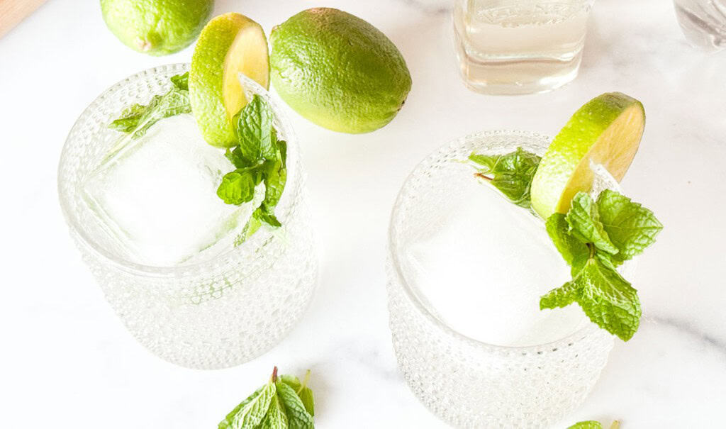 Two clear glasses filled with ice, mint sprigs, and lime slices sit on a white surface, with extra limes and mint nearby.