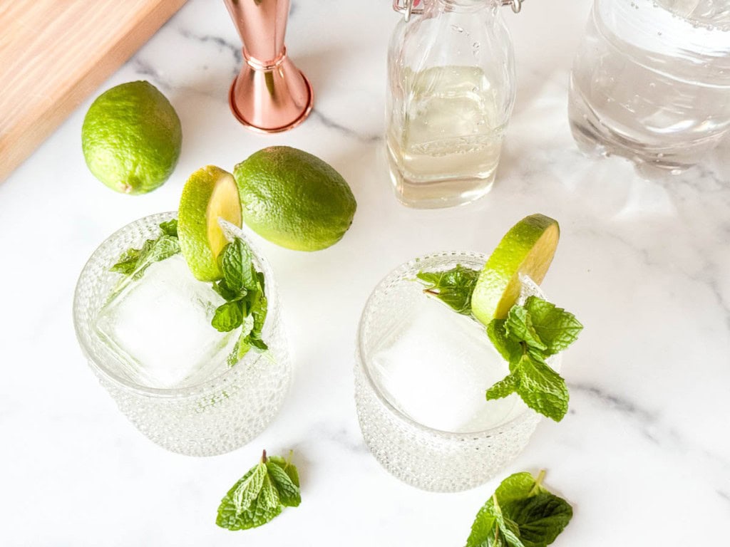 Two glasses with ice, lime slices, and mint leaves on a marble surface, alongside a bottle, a jigger, whole limes, and scattered mint.