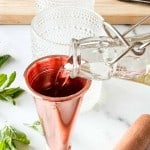 A clear liquid is being poured from a glass bottle into a copper jigger, with mint leaves, a muddler, and textured glassware nearby on a marble surface.