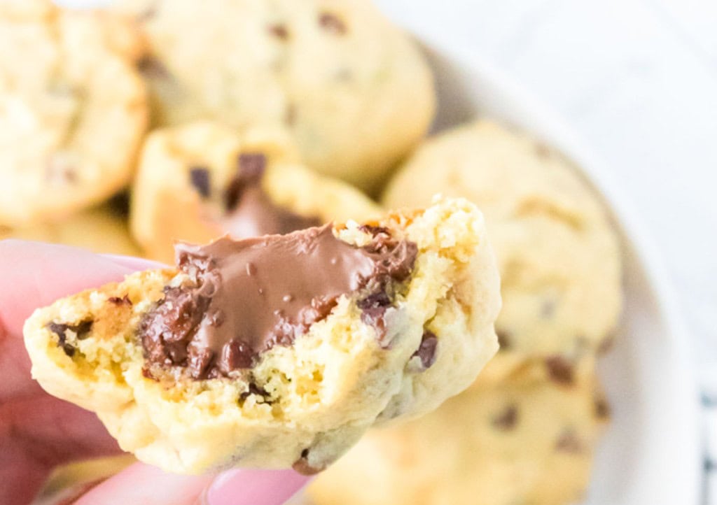 A close-up of a hand holding a chocolate chip cookie with a gooey chocolate filling and a bite taken out, with more cookies in the background.