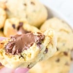 A close-up of a hand holding a chocolate chip cookie with a gooey chocolate filling and a bite taken out, with more cookies in the background.