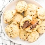 A plate of chocolate chip cookies, with one cookie broken in half to reveal a melted chocolate filling, placed on a white surface next to a checkered cloth.